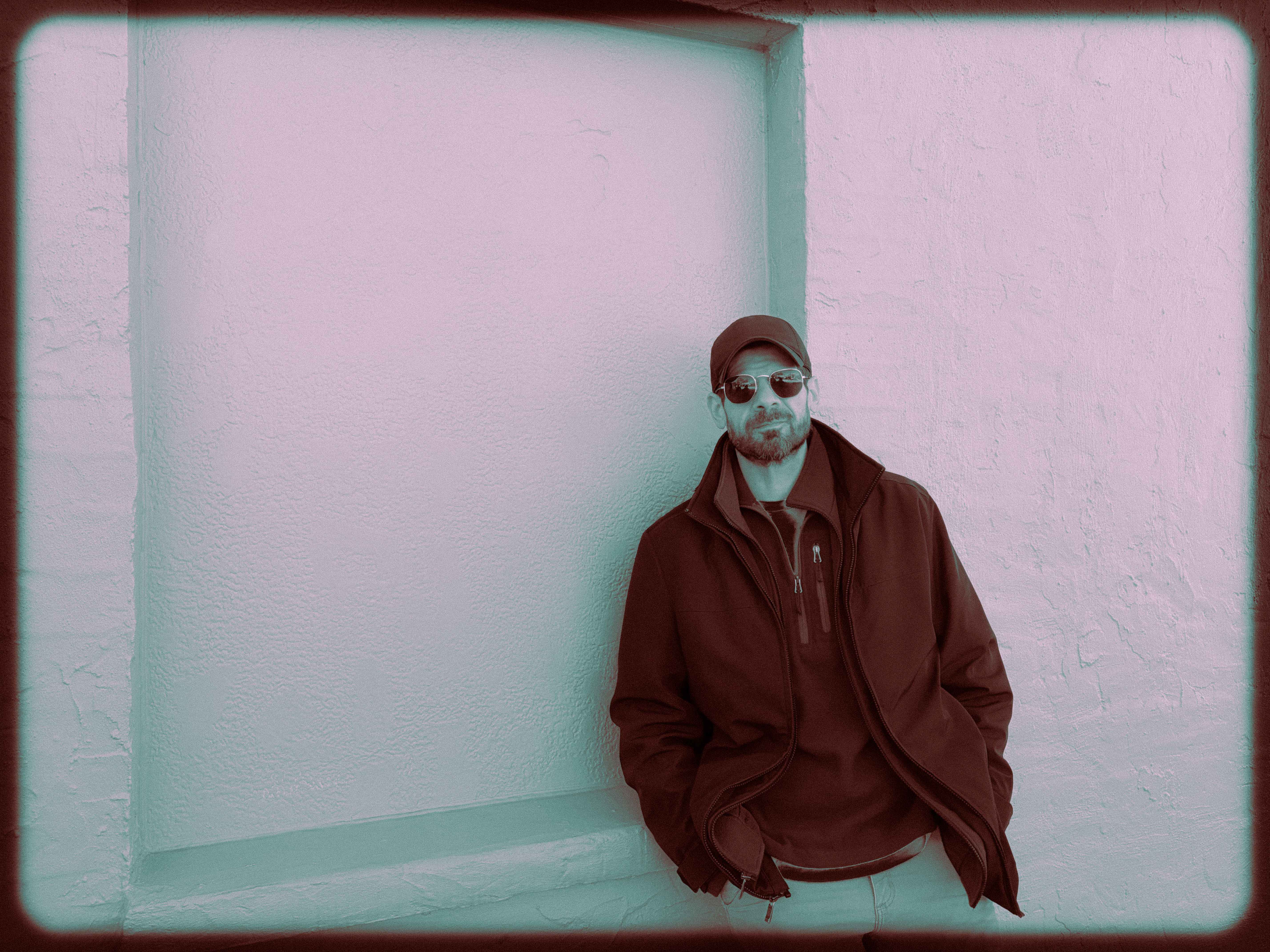 Michael Stakelum in a cap and aviator sunglasses, leaning against a textured stucco wall